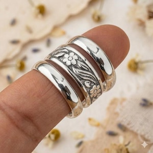 Könnte beinhalten: Ein Set aus drei gestapelten Ringen aus poliertem Silber. Der mittlere Ring zeigt ein geprägtes Blumenmuster mit kleinen Blüten und Blättern, während die beiden äußeren Ringe eine glatte, schlichte und abgerundete Oberfläche mit glänzendem Finish aufweisen.