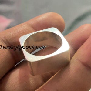 Puede incluir: Un anillo cuadrado de plata con un centro hueco. El anillo tiene una superficie lisa y pulida y un diseño geométrico moderno. El texto "Jewellrybyhandmade" es visible en la imagen.