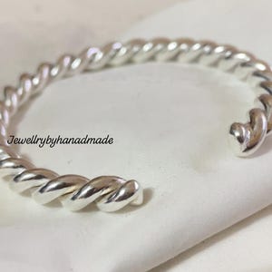 May include: A silver-colored cuff bracelet with a twisted rope design. The open-ended bracelet is made of a shiny metal and rests on a white surface. The text "Jewellrybyhandmade" is visible on the surface.
