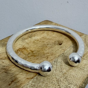 May include: A silver-colored cuff bracelet with a hammered texture and two rounded end pieces. The bracelet is open and rests on a wooden surface.
