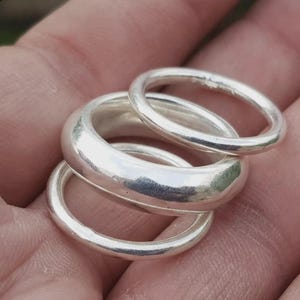 May include: A set of three polished silver-toned rings stacked together. The central ring features a thick, rounded band design, while the two outer rings are thinner and have a smooth, circular profile with a reflective metallic finish.