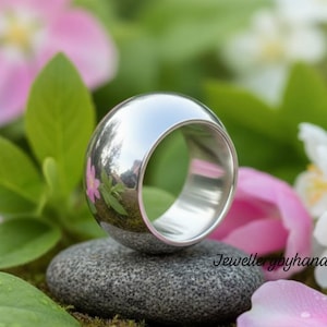 以下が含まれることがあります： 丸みを帯びたドーム型のトップが付いた、磨かれたシルバーリング。グレーの石の上に置かれています。リングは周囲の緑とピンクの花を反射しています。大胆でモダンなジュエリーです。