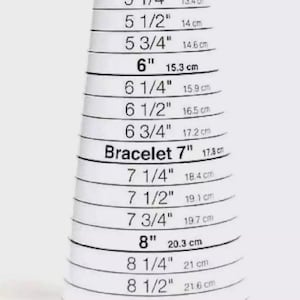 Może przedstawiać: Biały miernik EZ Bracelet Mini z czarnym tekstem wskazującym rozmiary bransoletki od 5 cali (12,7 cm) do 8 3/4 cala (22,2 cm). Każdy rozmiar jest oznaczony odpowiednikiem w calach i centymetrach.