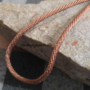 Puede incluir: Un collar de cadena de color cobre con un diseño tejido. El collar se muestra sobre una superficie de piedra gris texturizada. El texto "Jewellrybyhandmade" es visible en la piedra.