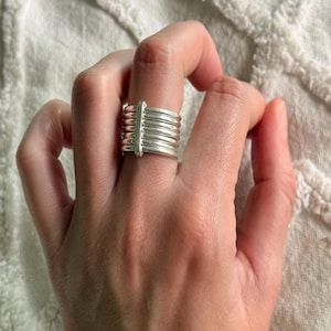 Puede incluir: Un anillo plateado con un diseño apilado. El anillo presenta múltiples bandas horizontales sujetas por una barra vertical. El anillo se lleva en un dedo, mostrando su brillo metálico y estilo moderno. El anillo es una pieza de joyería.