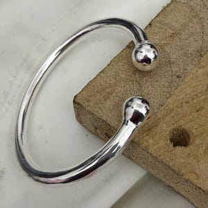 May include: A silver open cuff bracelet with a smooth, rounded design. The bracelet has a spherical ball at each open end. The jewellery is displayed on a wooden surface.