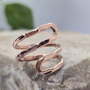 Peut inclure: Une bague de couleur cuivre présentant un design ouvert avec deux bandes qui se chevauchent. La surface métallique possède une finition martelée et texturée, créant une série de légères indentations le long du fil métallique en spirale.