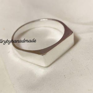 May include: A silver ring with a rectangular face. The ring is made of a shiny, reflective metal and is set against a white background. The text "Jewellrybyhanadmade" is in the upper left corner.
