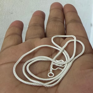 Puede incluir: Un collar de cadena plateada con un diseño texturizado en forma de cuerda. El collar se muestra en una mano, mostrando sus intrincados detalles y el cierre de cierre. La cadena está hecha de un metal brillante y reflectante.