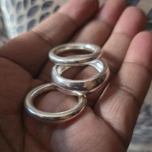Może przedstawiać: Trzy srebrne pierścionki o różnej szerokości są prezentowane w dłoni. Pierścionki mają gładkie, wypolerowane wykończenie, odbijające światło. Pierścionki są prawdopodobnie wykonane z metalu i mają prosty, klasyczny design.