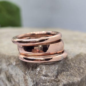 Puede incluir: Una pila de tres anillos pulidos en tono cobre. Los anillos superior e inferior son delgados y redondeados, mientras que el anillo central es más ancho con una superficie plana. El acabado metálico refleja la luz, mostrando una textura lisa.