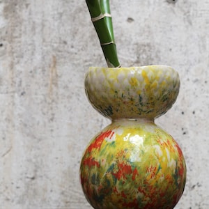 May include: A handcrafted ceramic vase with a unique hourglass shape. The vase features a vibrant, speckled design in yellow, red, and green. Two stalks of green bamboo are placed inside the vase. The vase is sitting on a wooden surface.