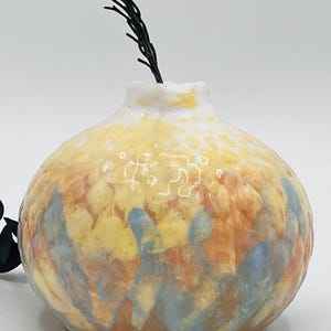 Puede incluir: Un pequeño jarrón redondo de cerámica con una abertura estrecha. El jarrón presenta una mezcla de tonos amarillos, naranjas y azules. Una ramita de follaje verde oscuro se extiende desde la parte superior. El jarrón tiene una superficie texturizada y un aspecto artesanal único.