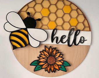 以下が含まれることがあります： 丸い木製の看板。ハチ、ハニカム、ヒマワリ、そして「hello」の文字が描かれています。ハチは黒と黄色の縞模様で、ヒマワリはオレンジと黄色です。家の装飾用です。