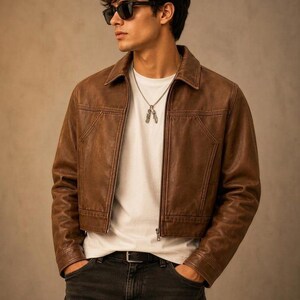May include: A brown leather jacket with a pointed collar and front zip fastening, worn over a plain white t-shirt. The outfit includes dark grey jeans with a black belt and a silver chain necklace with two small pendant charms.