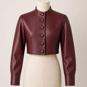 May include: A cropped jacket in a deep burgundy colour, featuring a high mandarin collar, long sleeves with wide cuffs, and a front button closure with five matching fabric-covered buttons. The jacket has a smooth, structured finish.