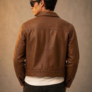 May include: A brown leather jacket with a classic collar, long sleeves, and visible vertical seam detailing on the back. The jacket is worn over a plain white t-shirt and dark-coloured jeans, showcasing a structured fit and a smooth texture.