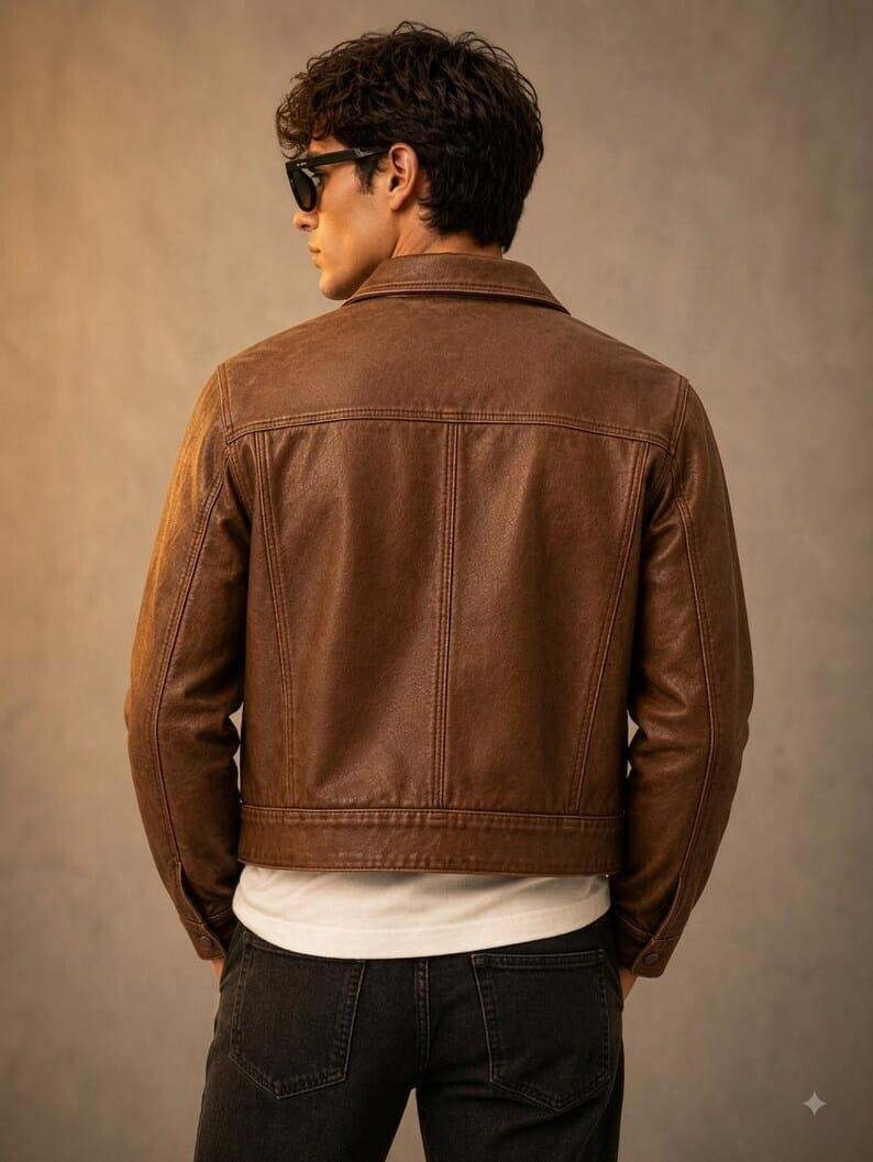 May include: A brown leather jacket with a classic collar, long sleeves, and visible vertical seam detailing on the back. The jacket is worn over a plain white t-shirt and dark-coloured jeans, showcasing a structured fit and a smooth texture.