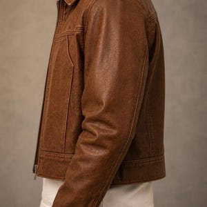 May include: A brown leather jacket featuring a classic collar, a full-length front zip, and long sleeves with buttoned cuffs. The jacket has a textured finish and is worn over a plain white t-shirt and dark-coloured trousers, showing a side profile.