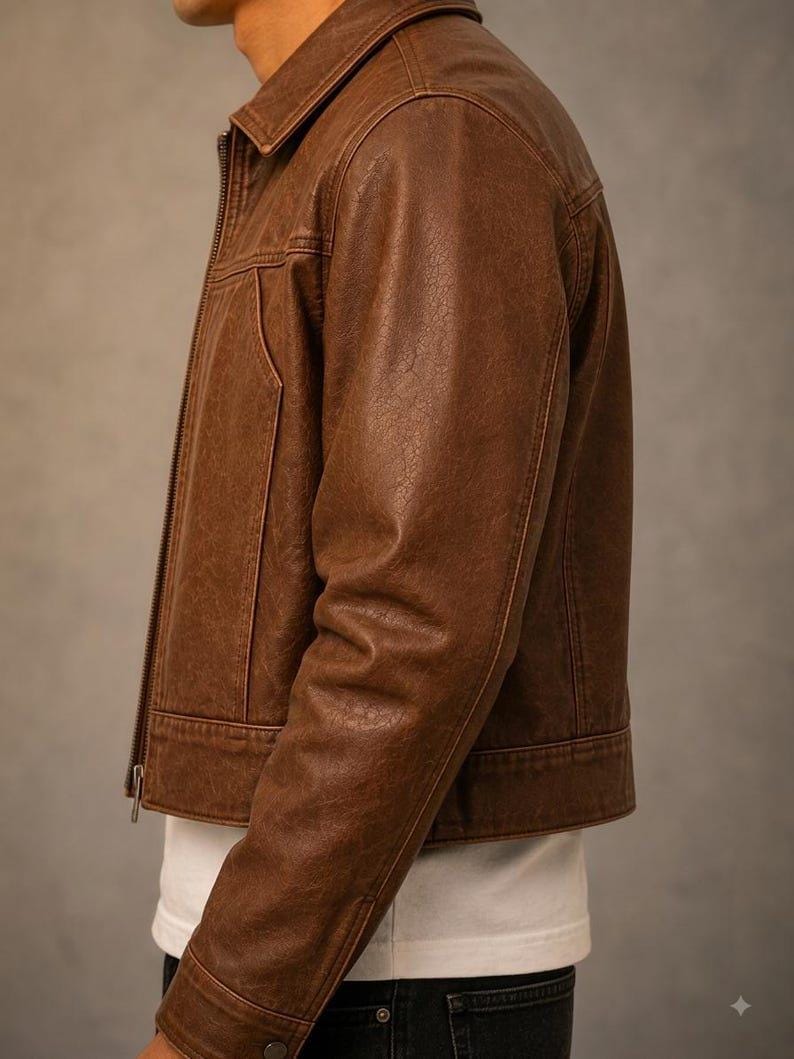 May include: A brown leather jacket featuring a classic collar, a full-length front zip, and long sleeves with buttoned cuffs. The jacket has a textured finish and is worn over a plain white t-shirt and dark-coloured trousers, showing a side profile.