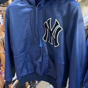 May include: A blue leather hoodie with a kangaroo pocket and a drawstring hood. The front features a large black and white embroidered logo. The jacket has long sleeves and a ribbed hem and cuffs.