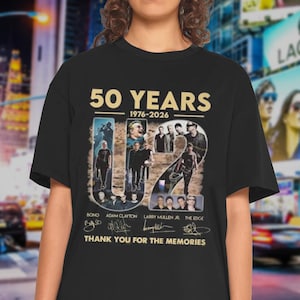 Op de afbeelding: Zwart t-shirt met een gouden afbeelding ter herdenking van 50 jaar, 1976-2026. Het ontwerp bevat afbeeldingen van een band, hun namen en de tekst "THANK YOU FOR THE MEMORIES".