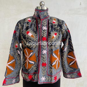 Può includere: Una giacca grigia con intricati ricami floreali nei toni dell'arancione, rosso e rosa chiaro. Il capo presenta un colletto alto, maniche lunghe e una chiusura frontale con profili rossi. Il testo Jaipurfabriccraft è visibile sul davanti.