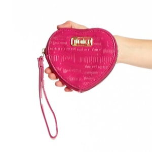 May include: A heart-shaped, cerise pink wallet with a wrist strap. The wallet has a zip closure and a gold-coloured metal plate with the word "Juicy" on it. The wallet is held in a hand against a white background.