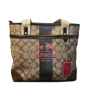 May include: A tan and brown monogram tote bag featuring a central dark brown vertical stripe, a red horse and carriage logo, and the text COACH LEATHERWARE EST. 1941. The bag includes cream-colored fabric handles, dark brown trim, and a red leather tag.