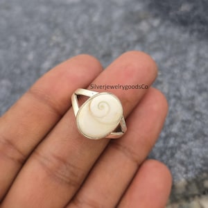 May include: A silver ring featuring an oval, off-white stone with a spiral design. The ring has a split band design. The text "SilverjewelrygoodsCo" is visible in the image.