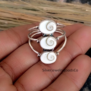 May include: Three silver rings with oval, white, shell-like inlays, each featuring a spiral design. The rings are stacked, creating a layered effect. The rings are set against a neutral background. The text "SilverjewelrygoodsCo" is visible.