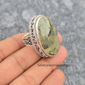 以下が含まれることがあります： 楕円形の淡い緑色の宝石が付いたシルバーリング。リングは、石の周りとバンドに装飾的なフィリグリーデザインが施されています。「SilverjewelrygoodsCo」というテキストが画像に表示されています。