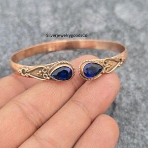 Pode incluir: Uma pulseira de punho aberta cor de cobre com duas pedras preciosas azuis escuras em forma de gota. A pulseira tem detalhes ornamentados perto das pedras preciosas. O texto "SilverjewelrygoodsCo" é visível.
