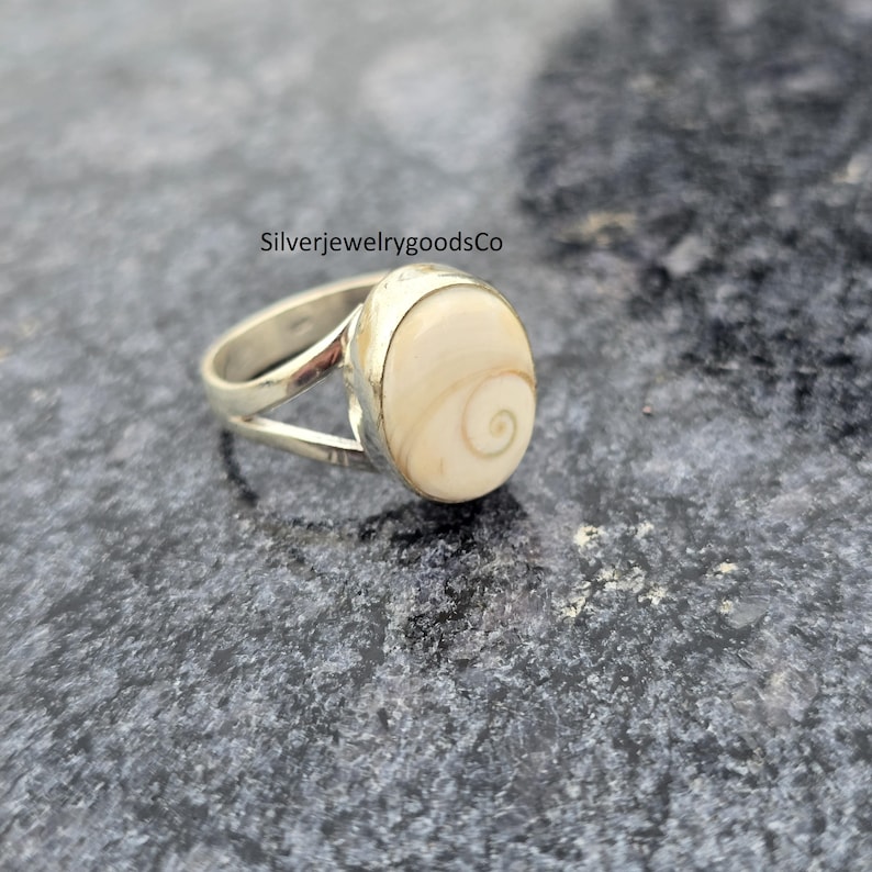 May include: A silver ring with a large, oval, cream-colored stone featuring a spiral design. The ring's band splits into two at the top, framing the stone. The text "SilverjewelrygoodsCo" is visible in the image.