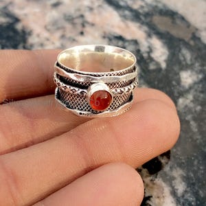 Può includere: Un anello spinner color argento con una gemma centrale, rotonda, arancione-rossa. L'anello ha un design strutturato con più fasce e un motivo intrecciato. L'anello è mostrato sul dito di una persona.