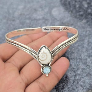 Pode incluir: Uma tiara prateada com uma concha branca em forma de gota e uma pequena pedra azul clara redonda. A tiara é segurada em uma mão. O texto "SilverjewelrygoodsCo" é visível na tiara.