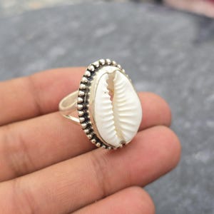 May include: A silver ring featuring a white cowrie shell centerpiece. The shell is oval-shaped and set within a beaded silver border. The ring's band is a simple, polished silver design, showcasing the natural beauty of the shell.