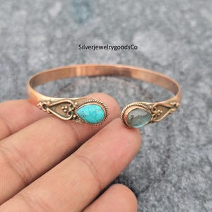 Puede incluir: Brazalete abierto de color cobre con piedras turquesas y azules en forma de lágrima. El brazalete tiene detalles ornamentados cerca de las piedras. El texto "SilverjewelrygoodsCo" es visible en el fondo.