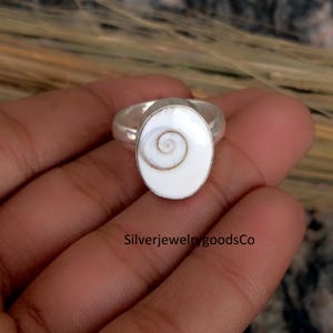 May include: A silver ring featuring an oval, white gemstone with a spiral design. The ring is set in a silver band. The image also includes the text "SilverjewelrygoodsCo".