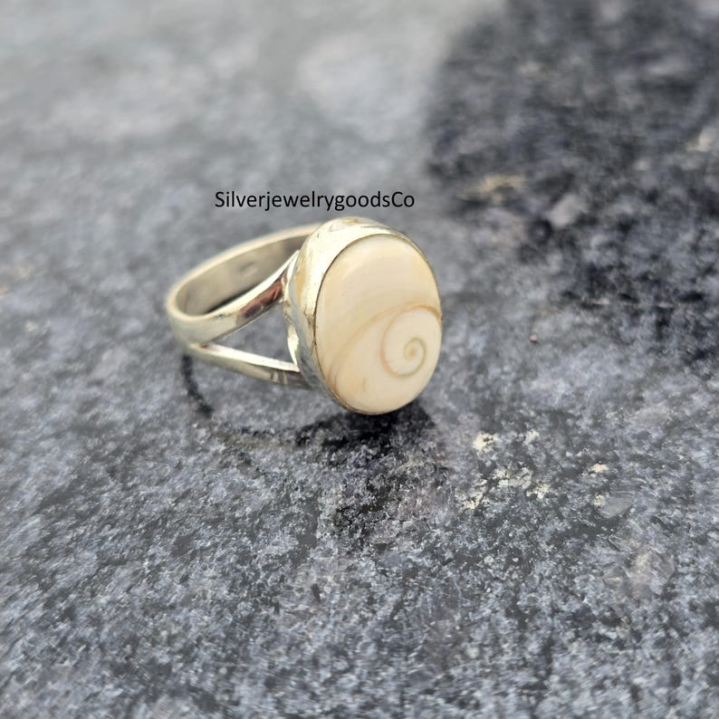 May include: A silver ring with a large, oval-shaped, cream-colored stone. The stone features a spiral design. The ring's band splits into two parts on either side of the stone. The text "SilverjewelrygoodsCo" is visible.
