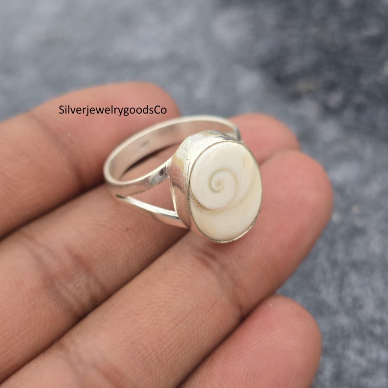 May include: A silver ring with a white, oval-shaped centerpiece featuring a spiral design. The ring has a split band design. The text "SilverjewelrygoodsCo" is visible in the image.