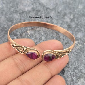 Op de afbeelding: Een goudkleurige open manchetarmband met twee druppelvormige, dieprode edelstenen. De armband heeft sierlijke, filigraan-achtige details bij de edelstenen. De tekst "SilverjewelrygoodsCo" is zichtbaar.