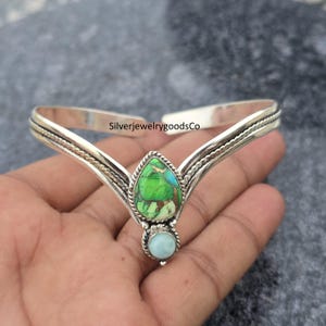 May include: A silver-toned crown-shaped bracelet with a central teardrop-shaped green and turquoise stone, and a smaller round blue stone below. The bracelet has a textured design and the text "SilverjewelrygoodsCo".