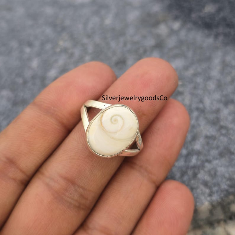 May include: A silver ring featuring an oval, off-white stone with a spiral design. The ring has a split band design. The text "SilverjewelrygoodsCo" is visible in the image.