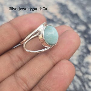 Puede incluir: Un anillo de plata con una piedra preciosa ovalada de color azul claro. El anillo tiene un diseño de caña dividida, con la banda dividida en dos hebras separadas que se curvan alrededor de la piedra preciosa. El anillo se muestra en una mano.