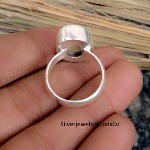 May include: A silver ring with a large, oval, light-colored stone. The ring band is smooth and rounded, and the stone is set in a bezel setting. The ring is held in a hand, with the text "SilverjewelrygoodsCo" visible.