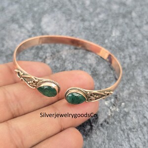 May include: Open cuff bracelet with a copper-colored band and two teardrop-shaped, emerald-green gemstones. The bracelet has ornate detailing near the gemstones. The text "SilverjewelrygoodsCo" is visible.