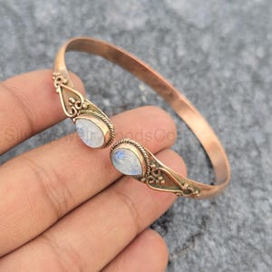Pode incluir: Uma pulseira de cobre com dois cabochões de pedra da lua em forma de lágrima nas extremidades abertas. A banda de metal apresenta detalhes intrincados de fio torcido e pequenos acentos esféricos em torno de cada engaste de pedra.
