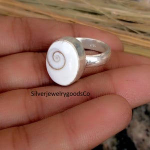 May include: A silver ring with a large, oval, white stone featuring a spiral design. The ring has a simple band and a bezel setting. The text "SilverjewelrygoodsCo" is visible at the bottom of the image.