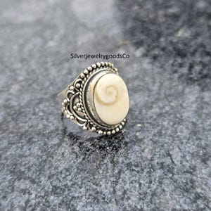 Puede incluir: Un anillo de plata con una piedra ovalada de color crema con un diseño en espiral. El anillo tiene un engaste ornamentado y detallado con un borde de cuentas y adornos decorativos. El texto "SilverjewelrygoodsCo" es visible.